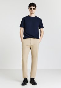 Mann in dunkelblauem Kurzarmhemd, beigen Hosen, schwarzen Schuhen und Sonnenbrille, der mit den Händen in den Taschen vor einem einfarbigen Hintergrund steht.