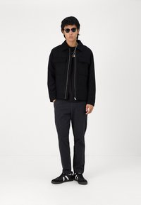 Veste zippée noire avec deux poches poitrine, portée sur une chemise noire. Pantalons gris texturés et baskets noires avec des rayures blanches. Lunettes de soleil rondes.