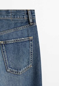 Détail de la poche arrière d’un jean en denim bleu avec passants de ceinture visibles et coutures sur fond blanc.