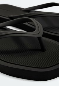 Gros plan de tongs en caoutchouc noir avec semelle texturée et brides en forme de Y sur fond blanc.