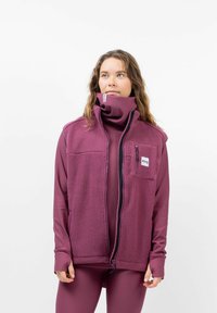 Fleece vest in een diepe pruimenkleur met een hoge kraag, rits aan de voorkant en zak; gedragen over een bijpassende aansluitende basislaag.