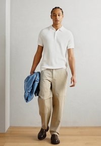 Wit geribd polo shirt, beige losse broek, en bruine schoenen. Spijkerjack in één hand. Effen achtergrond, houten vloer.