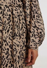 Vestido beige con estampado animal negro, con un cinturón atado en la cintura, mangas largas abullonadas y una tela suave y texturizada.