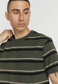 Camiseta de manga corta a rayas en verde oliva con rayas horizontales negras y beiges, con cuello redondo y una textura de tela suave.