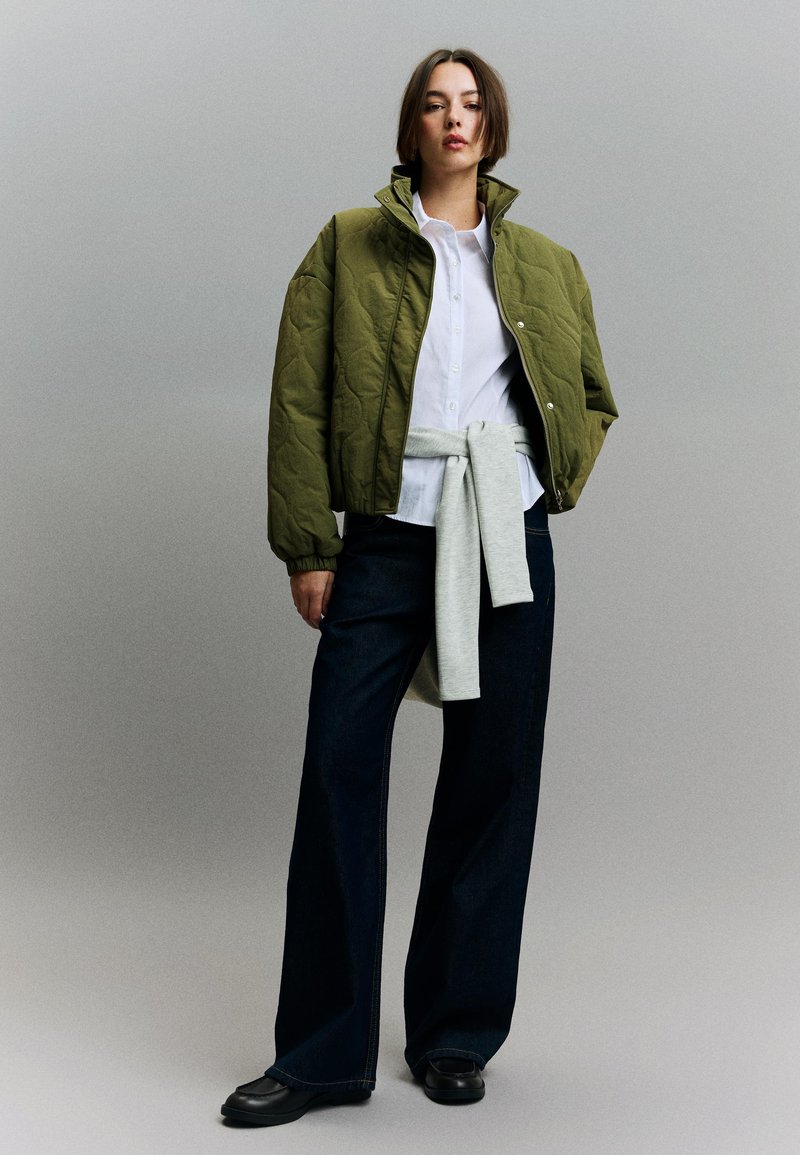 Kvinde iført olivengrøn vatteret jakke over hvid skjorte med grå sweater bundet om livet, mørke vidde-ben jeans og sorte sko mod ensfarvet baggrund.