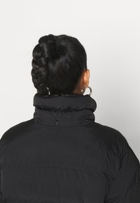 Chaqueta de plumas negra con un alto cuello y botones de presión, con un acabado texturizado. Cabello recogido en un moño trenzado.