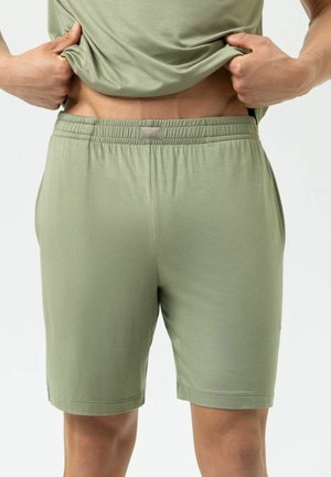 Person trägt hellgrüne Shorts mit elastischem Bund und hebt passendes Shirt an, um den unteren Torso vor hellem Hintergrund zu zeigen.
