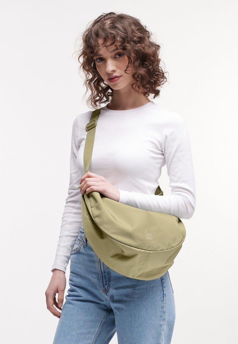 Jeune femme aux cheveux bouclés portant une chemise blanche à manches longues et un jean bleu, tenant un grand sac bandoulière vert olive.