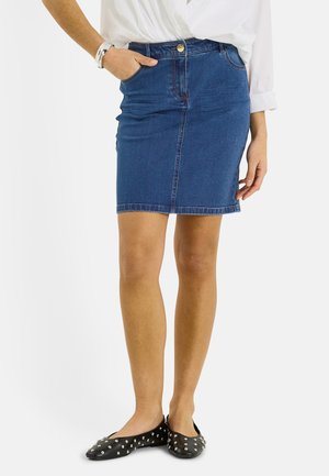 Mujer vistiendo una falda vaquera azul con bolsillos, blusa blanca con mangas arremangadas, pulseras plateadas y zapatos planos negros con tachuelas, de pie.