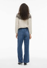 Bež pleteni pulover s uzorkom u smeđoj boji, uparen s trapericama širokih nogavica visokog struka u plavoj boji. Tekstura izgleda mekano i ležerno.