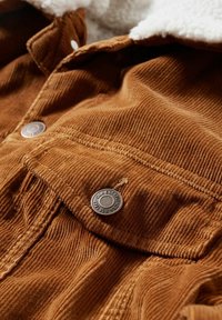 Brauner Cordstoff mit einer strukturierten Oberfläche, ausgestattet mit silbernen Metallknöpfen und einem weichen weißen Futter, das am Kragen sichtbar ist.