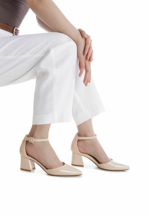 Tacones de charol beige con punta en punta y correa en el tobillo. Tacón grueso y translúcido que añade estabilidad. Llevados con pantalones blancos de pierna ancha.