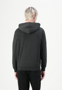 Personne avec des cheveux blonds courts portant un sweat à capuche gris foncé et un pantalon noir, de dos devant un fond blanc uni.