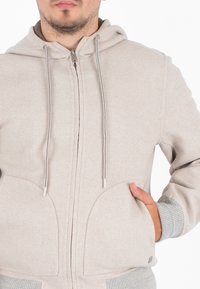 Felpa con zip color beige chiaro, dotata di cappuccio regolabile con cordino, tasche frontali e polsini a costine grigi. Tessuto morbido, design semplice e minimi dettagli.
