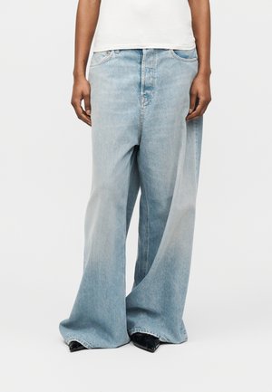 Άτομο που φοράει ανοιχτό μπλε τζιν με φαρδιά μπατζάκια και μαύρα μυτερά παπούτσια, στέκεται με τα χέρια χαλαρά στα πλάγια, μπροστά σε απλό φόντο.