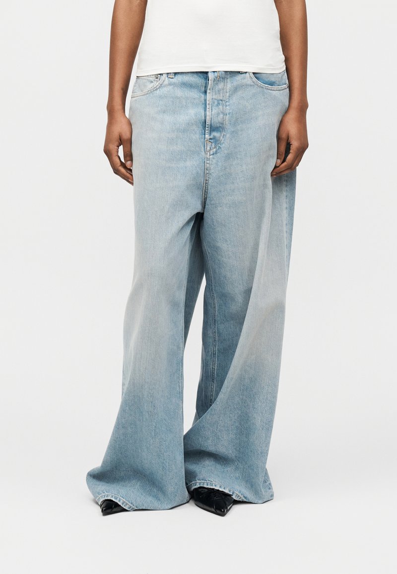 Άτομο που φοράει ανοιχτό μπλε τζιν με φαρδιά μπατζάκια και μαύρα μυτερά παπούτσια, στέκεται με τα χέρια χαλαρά στα πλάγια, μπροστά σε απλό φόντο.