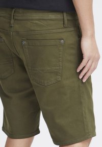 Olivgrüne Shorts aus robustem Stoff, mit zwei Gesäßtaschen, die sichtbare Nähte und Metallknopfakzente aufweisen.