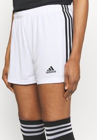 En person som bär vita Adidas shorts med svarta ränder och svarta knähöga strumpor med vita ränder, stående mot en neutral bakgrund.