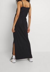 Robe maxi noire à fines bretelles, taille froncée et fente latérale ; portée avec des baskets blanches et grises pour un look décontracté.