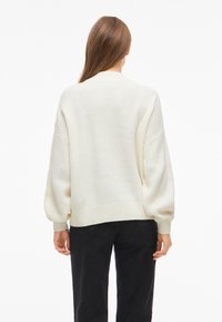 Frau mit langen braunen Haaren, die einen lockeren weißen gerippten Pullover und schwarze Hosen trägt, von hinten vor einem schlichten weißen Hintergrund gezeigt.