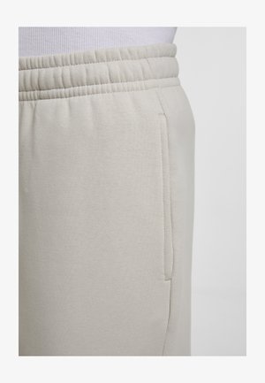 Close-up van beige joggingbroek met een elastische tailleband en zijzak, gedragen met een wit geribd shirt.