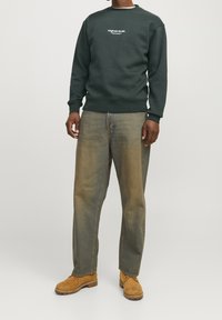 Uomo che indossa una felpa verde scuro con la scritta "Originals Studio", jeans larghi sbiaditi e stivali di suede beige appoggiato contro uno sfondo bianco.