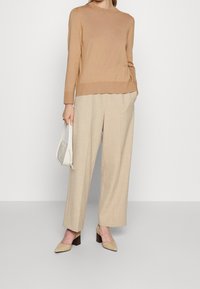 Kvinna som bär beige vida byxor, camelfärgad långärmad tröja, beige klackskor med blockklack och håller i en vit handväska.