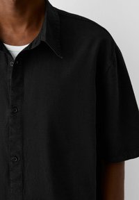 Detailní záběr na osobu v černé košili s krátkým rukávem s knoflíčky, která má na sobě bílou tričko s kulatým výstřihem.