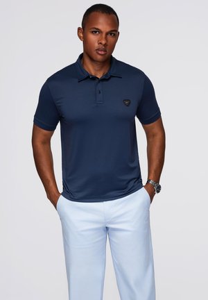 Ein Mann trägt ein navyblaues kurzärmliges Polo-Shirt und hellblaue Hosen, steht mit den Händen in den Taschen vor einem einfarbigen Hintergrund.