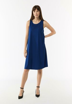Blaues ärmelloses Kleid mit runder Ausschnitt, fließendem Design und seitlichen Taschen, kombiniert mit schwarzen Ankle-strap-Pumps vor einem schlichten Hintergrund.