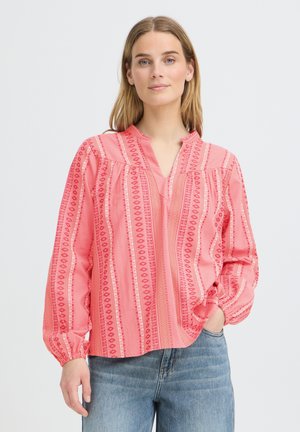Frau mit schulterlangem blondem Haar trägt eine pink gemusterte Langarmbluse und blaue Jeans, steht mit einer Hand in der Tasche.
