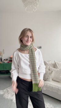 Eșarfă cu dungi verzi și crem, dintr-o textură tricotată, drapată peste un top alb cu mâneci lungi; pantaloni negri vizibili dedesubt.