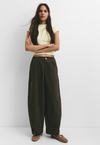 Geel geribde crop top, olijfgroene wijde broek met ritszakken en bruine instap schoenen met metalen accenten. Model staat met gekruiste armen.