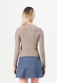 Femme aux longs cheveux blonds portant un pull côtelé beige et une jupe bleu forme trapèze, debout de dos sur un fond blanc uni.