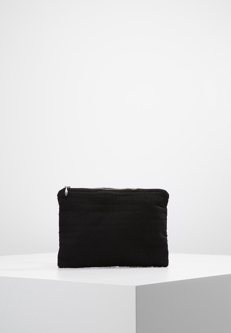Pochette rectangulaire en tissu noir avec une fermeture éclair sur le dessus, placée sur une surface angulaire blanche sur un fond gris clair uni.