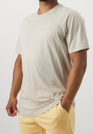 Homme portant un t-shirt beige Puma avec logo vertical sur la poitrine et pantalon jaune clair, main dans la poche sur fond uni.