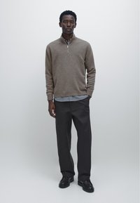 Brauner Strickpullover mit halbem Reißverschluss über einem hellblauen Hemd, kombiniert mit dunkler Hose und schwarzen Schuhen, stehend vor neutralem Hintergrund.