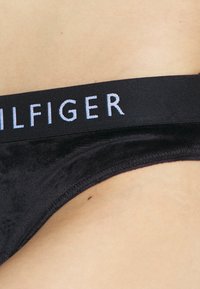 Svart stringtrosor med en mjuk textur, med en slank midjeband som visar "TOMMY HILFIGER" i ljusblå bokstäver.