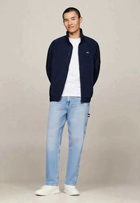 Casaco de ziper azul-marinho sobre uma t-shirt branca, combinado com jeans azul-claro e ténis brancos. Design simples, com detalhes de costura vis�íveis.