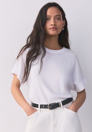 Kobieta o długich ciemnych włosach nosi białą koszulkę wpuszczoną w białe spodnie z czarnym paskiem, ręce w kieszeniach, na jednolitym jasnym tle.