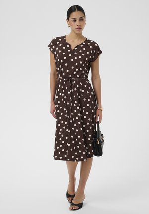 Femme debout portant une robe midi marron à pois blancs, tenant un sac à main noir et portant des talons ouverts noirs.