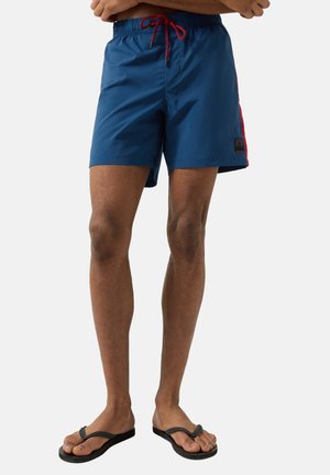 Mann trägt blaue Badeshorts mit roten Seitenstreifen und rotem Kordelzug, steht mit verschränkten Armen und trägt schwarze Flip-Flops vor grauem Hintergrund.