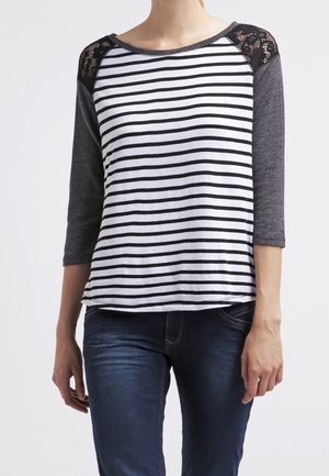 Femme portant une chemise rayée blanche et noire avec des épaules en dentelle gris foncé et des manches trois-quarts, associée à un jean bleu foncé.