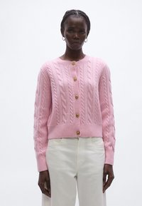 Rózsaszín kötött kardigán bordázott szegéllyel és mandzsettával, arany gombokkal az elején. Fehér nadrág fölött viselve.