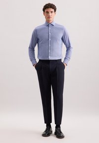 Uomo in piedi con le mani in tasca, che indossa una camicia a bottoni blu con motivo, pantaloni neri, calzini neri e scarpe eleganti nere su uno sfondo chiaro.
