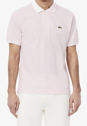 Homme portant un polo rose clair à rayures avec un col blanc et un logo crocodile vert, associé à un pantalon blanc.