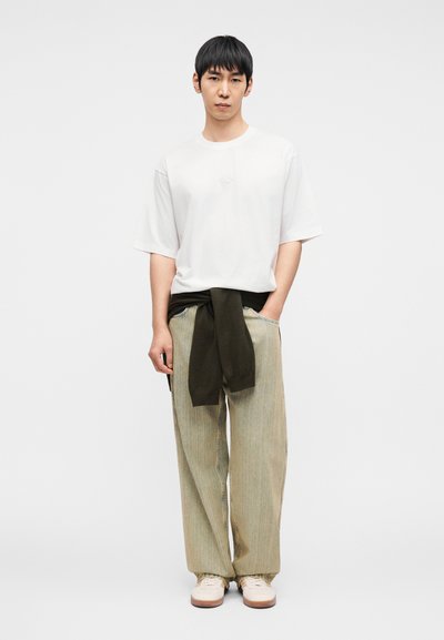 Λευκό T-shirt με κοντά μανίκια, πράσινο πλεκτό πουλόβερ δεμένο γύρω από τη μέση, ριγέ μπεζ παντελόνι με φαρδιά πόδια και κρεμ αθλητικά παπούτσια.