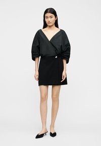 Γυναίκα που στέκεται, φοράει μαύρη μπλούζα με βαθύ V και φουσκωτά μανίκια, μαύρη μίνι φούστα με κουμπιά στο πλάι και μαύρες μυτερές γόβες.