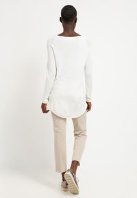 Maglione bianco a maniche lunghe con scollatura ampia, orlo curvo e dettagli a coste, abbinato a pantaloni slim-fit beige chiaro e calzature.
