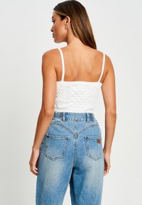 Top corto blanco de punto con tirantes finos, que presenta un patrón texturizado. Combinado con unos pantalones vaqueros de denim azul de tiro alto, que exhiben bolsillos traseros.